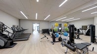 This fitness facility is designed to support a comprehensive workout experience within a clean, modern environment. The space features light wood-style flooring and a drop ceiling fitted with long, linear LED lighting panels that cast bright, even illumination throughout the room. Neutral grey walls contribute to a calm, focused atmosphere.

Along one side of the room, a full-length mirror wall visually expands the space and allows users to monitor their form during exercise. A row of treadmills lines the opposite wall, providing dedicated cardio space. The central floor area is home to a stationary bike and a multi-function cable resistance machine, offering versatile strength and conditioning options.

A dumbbell rack with a full range of free weights is positioned mid-room, accompanied by a flat utility bench. Colorful stability and medicine balls are displayed on a rack near the mirror wall, adding a functional training element. A doorway at the far end of the room leads to an additional area, suggesting the facility extends beyond the main floor space. Overall, the gym is well-organized, brightly lit, and equipped to accommodate a variety of fitness goals.