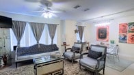 This single-floor space exudes a refined, eclectic charm, blending classic French-inspired design with contemporary decorative accents. The main living area serves as the focal point, showcasing a coordinated seating arrangement that includes an ornate sofa and a pair of carved, silver-toned armchairs, all upholstered in rich patterned fabric with coordinating throw pillows. A distinctive vintage steamer trunk repurposed as a glass-top coffee table anchors the seating area, adding a bold, character-filled touch to the room.

The space is bathed in natural light, courtesy of multiple windows dressed with elegant gray silk-style drapes over white blinds, creating a layered, polished window treatment throughout. A ceiling fan with integrated lighting provides both comfort and illumination overhead, while a sparkling chandelier visible in the corner adds a glamorous flourish.

The walls are kept in a clean, neutral tone that allows the bold patterned area rug and a striking piece of modern wall art to stand out as visual highlights. Decorative accents — including a star ornament, a side table lamp, and carefully placed accessories — complete the thoughtfully curated aesthetic. Overall, the space combines warmth, elegance, and personality in a well-appointed single-level layout.