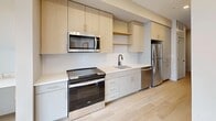 This efficiently designed one-bedroom, one-bathroom residence offers a smart, modern layout with clean lines and a bright, neutral palette throughout. Light hardwood flooring flows continuously from the entry corridor through the main living areas, complemented by recessed lighting and crisp white millwork with matte black hardware accents.

The kitchen is outfitted with two-tone cabinetry — light wood uppers paired with soft grey lowers — white quartz countertops, a subway tile backsplash, and a full suite of stainless steel appliances including a range, microwave, dishwasher, and refrigerator. Open shelving provides both storage and display space, while a built-in desk nook adjacent to the kitchen adds functional versatility.

The combined living area is filled with natural light from large floor-to-ceiling windows and a sliding glass door that opens to a private balcony with urban views. The bedroom is a calm, neutral retreat featuring a generous walk-in closet with built-in shelving and hanging rods. The bathroom includes a soaking tub and shower combination, a wide vanity with white cabinetry, a large mirror, and matte black fixtures. A well-proportioned hallway with sliding barn doors connects all areas of the residence seamlessly.