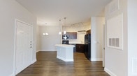Located at 2030 Wildts Battery Blvd in Johns Island, South Carolina, this single-floor residence offers a thoughtfully designed open-concept layout that seamlessly connects the living and kitchen areas. Rich, dark hardwood-style flooring runs throughout, providing a warm and cohesive foundation for the space.

The kitchen is a standout feature, showcasing dark espresso cabinetry that contrasts elegantly with a crisp white center island, ideal for meal preparation or casual dining. Black appliances, including a range and a full-size refrigerator, complement the cabinetry's rich tones. Track lighting above the cooking area ensures the workspace is well-lit, while stylish pendant lights suspended above the island add a decorative touch. A sink is positioned along the perimeter cabinetry, offering a functional layout for cooking and cleanup.

The adjacent living area benefits from the open sightlines, creating an inviting and spacious atmosphere. An elegant chandelier overhead adds a refined layer of lighting and character to the shared space. Neutral-toned walls and white trim throughout keep the interior feeling bright, clean, and versatile — ready to accommodate a range of interior styles. The single-floor plan ensures easy accessibility and a comfortable, connected flow between all areas of the home.