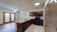 This single-floor unit offers a well-designed open-concept layout that seamlessly connects the kitchen and living areas. The kitchen serves as a central hub, featuring rich mahogany-toned cabinetry, granite-style countertops, and a peninsula island with a double sink and built-in dishwasher. Black appliances, including a range, microwave, and refrigerator, complement the dark cabinetry for a cohesive aesthetic. Pendant lighting hangs above the peninsula, adding a refined touch to the space. Tile flooring in the kitchen transitions into warm, dark hardwood flooring throughout the living and bedroom areas, creating a clear and attractive visual distinction between spaces. The living area is generously proportioned, illuminated by natural light from French doors and adjacent windows fitted with blinds, and finished with a ceiling fan overhead. The bedroom is a comfortable, well-lit space featuring two windows with blinds and its own ceiling fan. The bathroom is equipped with a dark-topped vanity, tile flooring, a wall-mounted mirror cabinet, and an accessible roll-in shower with grab bars, offering both style and practicality. Overall, the unit presents a clean, functional, and inviting living environment.
