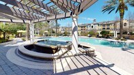 This single-story amenity space is defined by its resort-inspired outdoor design, combining elegant architectural details with well-appointed recreational features. At the heart of the outdoor area sits a generously sized swimming pool, complemented by an adjacent spa with decorative stone cladding and active water features. A substantial pergola structure, supported by classic white columns, provides partial shade over the spa area while framing views of the surrounding landscaped grounds and community buildings beyond.

The hardscape throughout features intricate interlocking pavers that extend across the pool deck, walkways, and covered porch areas, lending a cohesive and refined aesthetic. Mature palm trees and carefully maintained shrubs and hedges punctuate the grounds, enhancing the tropical ambiance.

The covered porch area showcases a combination of green lap siding and stacked stone masonry, with large multi-pane windows and French doors that allow natural light to flow through. Traditional wall-mounted lanterns and a ceiling fan add functional charm to the porch, which is furnished with Adirondack rocking chairs ideal for relaxed outdoor seating. Pergola-lined walkways connect the various zones, creating a seamless, welcoming environment suited for leisure and community gathering.