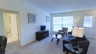 This two-floor residence offers a well-organized layout with clean, neutral interiors throughout. The living room serves as the central gathering space, featuring plush wall-to-wall carpeting in a warm beige tone that adds comfort underfoot. Smooth, light-colored walls provide a bright and airy backdrop, complemented by a large picture window that allows generous natural light to fill the room and offers views of the surrounding neighborhood. The living area is furnished with a dark sofa and a patterned accent chair arranged around a sleek glass-top coffee table, creating a balanced and functional seating arrangement. A dark wood media console houses a flat-screen television along one wall, while framed artwork adds a decorative touch to the open wall space. A partial view of an adjacent area, likely a kitchen or dining space, is visible at the edge of the room, suggesting an open or semi-open floor plan. The overall ambiance of the space is clean, understated, and practical, with a neutral color palette that lends itself well to personalization. The two-floor design provides additional living or sleeping space, offering versatility for a variety of lifestyle needs.