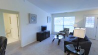 This two-floor residence offers a well-organized layout with clean, neutral interiors throughout. The living room serves as the central gathering space, featuring plush wall-to-wall carpeting in a warm beige tone that adds comfort underfoot. Smooth, light-colored walls provide a bright and airy backdrop, complemented by a large picture window that allows generous natural light to fill the room and offers views of the surrounding neighborhood. The living area is furnished with a dark sofa and a patterned accent chair arranged around a sleek glass-top coffee table, creating a balanced and functional seating arrangement. A dark wood media console houses a flat-screen television along one wall, while framed artwork adds a decorative touch to the open wall space. A partial view of an adjacent area, likely a kitchen or dining space, is visible at the edge of the room, suggesting an open or semi-open floor plan. The overall ambiance of the space is clean, understated, and practical, with a neutral color palette that lends itself well to personalization. The two-floor design provides additional living or sleeping space, offering versatility for a variety of lifestyle needs.