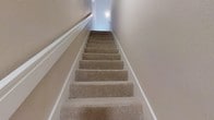 This 1-bedroom, 1-bathroom residence spans three floors, offering a thoughtfully distributed layout that maximizes the use of its square footage. The space is connected by a carpeted staircase, which features neutral beige tones and crisp white trim along the walls, creating a clean and cohesive aesthetic throughout the vertical circulation of the home. The staircase is well-lit with wall-mounted lighting, ensuring safe and comfortable movement between levels. The neutral color palette of the walls and flooring throughout the stairwell reflects an overall design sensibility that is understated and practical. The multi-floor configuration allows for a natural separation of living, sleeping, and utility spaces across the three levels, lending the residence a sense of organization that single-floor units often lack. The soft carpeting on the stairs adds warmth and sound dampening to the interior environment. Overall, the space presents a functional and comfortable home suited for those who appreciate a structured, multi-level living arrangement within a modest and well-maintained footprint.