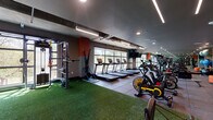 This expansive fitness center is designed to accommodate a wide range of training styles within a thoughtfully organized, single-level layout. The space is defined by its industrial-modern aesthetic, featuring exposed concrete ceilings, bold orange accent walls, and a combination of recessed and linear LED lighting that ensures the entire area is well-illuminated throughout.

A row of Life Fitness treadmills lines a floor-to-ceiling window wall, providing users with direct views of the outdoors and an abundance of natural light during workouts. Adjacent to the cardio row, a lineup of cycling bikes offers additional cardiovascular options. Large mirrors along one wall visually expand the space and support proper form during strength training.

A generously sized turf zone serves as the centerpiece of the functional training area, equipped with battle ropes, weighted sleds, tires, medicine balls, a freestanding punching bag, and a multi-use cable rig. This zone transitions seamlessly into a dedicated strength section featuring dumbbells, benches, and cable machines.

A separate glass-enclosed studio room with wood-look flooring and sleek linear ceiling lighting provides a quieter space for focused training or stretching. A wall-mounted mat and water station add practical convenience throughout the facility.