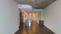 This single-floor space showcases a compelling blend of industrial and contemporary design. The open-plan layout connects the kitchen and living areas, creating a cohesive, flowing environment. A defining architectural feature is the curved drywall ceiling detail that contrasts strikingly with the raw, exposed concrete ceiling beyond it, adding visual depth and character to the space.

Dark hardwood flooring runs throughout, providing a warm, grounding element that complements the industrial aesthetic. The kitchen is centrally anchored by a peninsula-style island with an integrated sink, offering both functional workspace and a natural visual divider between zones. Stainless steel appliances, including a refrigerator and oven, line the kitchen wall, while dark cabinetry adds a sophisticated touch. Pendant lighting above the island provides focused illumination.

An accent wall featuring exposed concrete or stone texture near the entryway further reinforces the industrial character of the space. The open sightlines from the main living area through to the entryway enhance the sense of spaciousness. Overall, the space delivers a modern urban ambiance defined by contrasting textures, clean lines, and thoughtful architectural detailing.