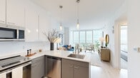 Located at 909 E Michigan St in Milwaukee, Wisconsin, this well-appointed one-bedroom, one-bathroom residence offers a thoughtfully designed layout with modern finishes and breathtaking views of Lake Michigan. The open-concept kitchen and living area is a highlight of the space, featuring two-tone cabinetry with crisp white upper cabinets and slate-gray lower cabinets, a clean white countertop, stainless steel appliances including a refrigerator, range, microwave, and dishwasher, and pendant lighting over the kitchen island. The living area flows seamlessly toward a dramatic wall of floor-to-ceiling windows, flooding the space with natural light and framing panoramic lake and cityscape views. Light hardwood flooring runs throughout the main living areas, lending warmth to the contemporary design. The bedroom continues the floor-to-ceiling window theme, offering serene waterfront views and plush carpeting underfoot. The bathroom is clean and modern, with gray cabinetry, a white countertop, a large mirror, and a full tub-shower combination with white tile surround. The hallway entry accommodates a stacked in-unit washer and dryer alongside a closet, maximizing functionality. The overall ambiance is bright, airy, and modern, with a neutral palette that complements the stunning outdoor scenery.