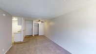 This single-floor residential space offers a straightforward and functional layout designed for comfortable everyday living. The main living area is finished with wall-to-wall carpeting in a warm neutral tone, providing a soft and inviting underfoot feel that extends throughout the space. A ceiling fan with an integrated light fixture is centrally positioned overhead, ensuring air circulation and ambient lighting across the room. The textured popcorn ceiling adds a classic, familiar character to the interior. Neutral-toned walls keep the space feeling clean and versatile, lending itself easily to a variety of furnishing styles. A built-in closet with open shelving provides convenient storage directly off the main living area, while a solid-panel entry door offers a clear and practical point of access. A partial view of an adjacent kitchen area is visible along the side wall, suggesting a connected and efficient floor plan. The overall ambiance is calm and understated, with a blank-canvas quality that makes it easy to personalize. The space is well-suited for those seeking a simple, low-maintenance living environment with essential features in a compact, single-level arrangement.