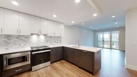 This thoughtfully designed single-floor residence showcases a clean, modern aesthetic with warm hardwood-style flooring and recessed lighting throughout. The open-concept layout seamlessly connects the living area to the kitchen, creating a fluid and functional living space.

The kitchen is a standout feature, outfitted with two-toned cabinetry — crisp white upper cabinets paired with dark lower cabinets — and a striking blue-accented island with a white quartz countertop and an undermount sink. A marble-style tile backsplash adds elegance, while stainless steel appliances, including a gas range, refrigerator, and built-in microwave, complete the chef-ready space.

The living area is bright and airy, with neutral walls and large sliding glass doors that open to a private balcony, inviting natural light and outdoor views. The bedroom is generously proportioned, featuring a large window that frames neighborhood views and fills the room with daylight.

The bathroom is finished with striking marble-look porcelain tile covering both the floors and the tub surround, creating a cohesive, spa-like atmosphere. A combination tub and shower with a handheld showerhead rounds out the refined finishes. Additional storage and in-unit laundry connections are also present, enhancing the overall practicality of this well-appointed residence.