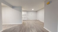 This single-floor residence offers a clean, neutral aesthetic throughout, with light gray walls and wood-look luxury vinyl plank flooring unifying the main living areas. The entry opens directly into a generously sized living room, characterized by a large window that admits abundant natural light and provides views of the surrounding outdoor environment. A wall-mounted A/C unit and baseboard heating ensure year-round climate comfort.

The kitchen is thoughtfully designed with white shaker-style cabinetry extending from floor to ceiling, complemented by light marble-patterned countertops, a stainless steel sink, and a dishwasher. Recessed lighting illuminates the workspace efficiently. An open pass-through layout connects the kitchen to an adjacent area, enhancing the overall sense of spaciousness.

The bedrooms feature plush wall-to-wall carpeting and individual windows overlooking greenery, with baseboard heating units beneath each window. The bathroom is finished with a full soaking tub and tub surround clad in marble-look tile, paired with clean white fixtures and wood-look flooring consistent with the rest of the unit. Overall, the space presents a modern, move-in-ready interior with a cohesive and functional design.