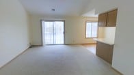 This single-floor space features an open-concept layout that combines a spacious living area with an adjoining kitchen. The living room is finished with light-colored carpeting that extends across the generous floor area, creating a soft, comfortable underfoot feel. A vaulted ceiling adds a sense of vertical openness and architectural interest, elevating the overall ambiance of the main living space.

Natural light is a defining feature of this space. A large sliding glass door dominates one wall of the living area, offering direct access to an outdoor deck and framing views of the surrounding exterior. A secondary window, trimmed in warm wood tones, provides additional light and ventilation. Together, these openings keep the interior bright and airy throughout the day.

Transitioning toward the kitchen, hardwood or laminate flooring replaces the carpet, providing a durable and easy-to-maintain surface. The kitchen area includes cabinetry finished in a warm brown tone and a light-colored countertop that wraps along the perimeter. A range hood is mounted above, indicating a dedicated cooking zone.

The neutral color palette of soft yellows and whites throughout the space creates a clean, versatile backdrop suitable for a variety of interior styles.