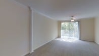 This single-floor space presents an open, flowing layout that connects the living area to the kitchen and beyond. The main living area is spacious and bright, highlighted by a large sliding glass door framed with vertical blinds that invites abundant natural light and provides views of greenery outdoors. A ceiling fan is centrally positioned overhead, and elegant crown molding lines the perimeter of the ceiling, adding a refined architectural detail. A decorative white column serves as a visual divider within the open floor plan while maintaining the sense of openness throughout.

Soft, neutral-toned carpet covers the floors, contributing to a warm and comfortable atmosphere. The walls are finished in a light cream tone, complementing the overall brightness of the space. Recessed and track lighting fixtures are installed throughout, providing adjustable illumination across various areas.

Toward the rear, the kitchen area features a breakfast bar or counter that opens toward the living space, enhancing the connectivity of the floor plan. A hallway leads to additional rooms, including a closeted space visible near the entry. The overall ambiance is clean, airy, and versatile, well-suited for a variety of living arrangements.