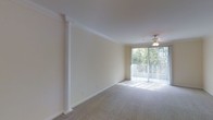 This single-floor space presents an open, flowing layout that connects the living area to the kitchen and beyond. The main living area is spacious and bright, highlighted by a large sliding glass door framed with vertical blinds that invites abundant natural light and provides views of greenery outdoors. A ceiling fan is centrally positioned overhead, and elegant crown molding lines the perimeter of the ceiling, adding a refined architectural detail. A decorative white column serves as a visual divider within the open floor plan while maintaining the sense of openness throughout.

Soft, neutral-toned carpet covers the floors, contributing to a warm and comfortable atmosphere. The walls are finished in a light cream tone, complementing the overall brightness of the space. Recessed and track lighting fixtures are installed throughout, providing adjustable illumination across various areas.

Toward the rear, the kitchen area features a breakfast bar or counter that opens toward the living space, enhancing the connectivity of the floor plan. A hallway leads to additional rooms, including a closeted space visible near the entry. The overall ambiance is clean, airy, and versatile, well-suited for a variety of living arrangements.