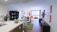 This single-floor residence offers a thoughtfully designed open-concept layout that seamlessly connects the kitchen, dining, and living areas into one cohesive and inviting space. Rich dark hardwood flooring runs throughout, grounding the interior with warmth and sophistication.

The kitchen features a sleek countertop peninsula that serves as both a functional workspace and a natural divider between the cooking and dining areas. A compact dining nook with a round table and modern chairs sits just beyond, creating an intimate setting for meals.

The living area is anchored by vibrant accent rugs and flows effortlessly toward a large floor-to-ceiling window that spans the far wall, bathing the space in abundant natural light and offering open urban views. The window's generous proportions enhance the sense of openness and connect the interior to the cityscape beyond.

Décor throughout strikes a balance between contemporary and eclectic, with a decorative wall mirror, open shelving units displaying curated accessories, and warm accent lighting provided by track fixtures overhead. The overall ambiance is bright, modern, and welcoming — ideal for both everyday living and entertaining.