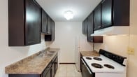 This single-story space features a thoughtfully designed galley-style kitchen that maximizes functionality within a compact, efficient layout. The kitchen runs parallel along two walls, creating a streamlined corridor that keeps everything within easy reach. Dark espresso-toned cabinetry lines both sides of the kitchen from floor to ceiling, providing generous storage space while adding a bold contrast against the neutral, light-colored walls. The upper cabinets extend high toward the textured ceiling, making excellent use of vertical space.

Laminate countertops in a warm, earthy tone run along both sides of the kitchen, offering ample workspace for meal preparation. On one side, a double-basin stainless steel sink is positioned beneath the cabinetry, accompanied by a standard faucet. The opposite counter houses a white electric coil-burner range with an overhead exhaust hood, keeping the cooking area well-ventilated. A white refrigerator anchors the far end of the kitchen. Light neutral tile flooring runs throughout, and a central flush-mount ceiling light ensures the space is well-illuminated. The overall aesthetic is clean, practical, and functional.