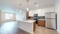 Located at 18000 W 97th St in Lenexa, Kansas, this well-appointed one-bedroom, one-bathroom residence offers a thoughtfully designed open-concept layout. The combined kitchen, dining, and living area serves as the heart of the home, showcasing a sleek kitchen with a central island featuring an undermount sink, crisp white upper cabinetry, warm wood-toned lower cabinets, a textured gray tile backsplash, and a full suite of stainless steel appliances including a refrigerator, range, and microwave. Elegant pendant lighting hangs above the island, adding a modern touch. Light wood-style flooring flows throughout the main living areas, complemented by crown molding for a refined finish. Large windows invite generous natural light into the living and dining spaces, offering pleasant outdoor views. The bedroom features plush carpeting and a window with exterior views, providing a comfortable retreat. Three closets offer ample storage throughout the unit. The bathroom is well-finished with wood-toned cabinetry, a white quartz-style countertop, an undermount sink, and a soaking tub framed by marble-look tile surround. Wood-style flooring continues into the bathroom, tying the design together cohesively.