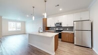 Located at 18000 W 97th St in Lenexa, Kansas, this well-appointed one-bedroom, one-bathroom residence offers a thoughtfully designed open-concept layout. The combined kitchen, dining, and living area serves as the heart of the home, showcasing a sleek kitchen with a central island featuring an undermount sink, crisp white upper cabinetry, warm wood-toned lower cabinets, a textured gray tile backsplash, and a full suite of stainless steel appliances including a refrigerator, range, and microwave. Elegant pendant lighting hangs above the island, adding a modern touch. Light wood-style flooring flows throughout the main living areas, complemented by crown molding for a refined finish. Large windows invite generous natural light into the living and dining spaces, offering pleasant outdoor views. The bedroom features plush carpeting and a window with exterior views, providing a comfortable retreat. Three closets offer ample storage throughout the unit. The bathroom is well-finished with wood-toned cabinetry, a white quartz-style countertop, an undermount sink, and a soaking tub framed by marble-look tile surround. Wood-style flooring continues into the bathroom, tying the design together cohesively.