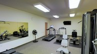 This fitness center is a dedicated, single-floor space designed to meet a variety of exercise needs. The room is bright and well-lit, featuring multiple recessed panel ceiling lights that illuminate the entire area evenly. The light-colored walls and speckled flooring contribute to a clean, functional atmosphere.

The cardio section is the centerpiece of the space, housing multiple treadmills equipped with individual display consoles, as well as an elliptical machine visible through a large wall mirror. The mirror not only adds a sense of depth to the room but also allows users to monitor their form during workouts. A standalone floor fan provides additional airflow and comfort during exercise.

Strength and flexibility training are also supported, with a weight bench and additional exercise equipment positioned throughout the space. A wall-mounted flat-screen television is prominently placed for entertainment during workouts.

A standard interior door provides access to an adjoining area or utility space, adding practical convenience. The overall layout is thoughtfully organized to allow comfortable movement between equipment. This fitness center offers a straightforward, no-frills environment well-suited for daily exercise routines.