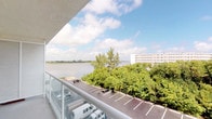 This single-floor space is distinguished by its private balcony, which serves as a standout architectural feature and a true highlight of the property. Framed by a sleek, modern railing, the balcony opens up to a commanding panoramic view of a tranquil waterway, lush tree canopy, and expansive open skies dotted with clouds. The elevated vantage point provides a sense of openness and connection to the natural surroundings, making it an ideal spot for relaxation or enjoying the outdoors. The waterfront scenery, combined with the lush greenery below, creates a peaceful and picturesque backdrop that can be enjoyed throughout the day. The balcony's clean, contemporary design complements the overall aesthetic of the space. While much of the interior is not fully visible in the available imagery, the presence of a well-appointed entryway and additional interior areas can be discerned, suggesting a thoughtfully laid-out floor plan. The space benefits from its elevated position, which not only enhances the waterfront views but also provides a sense of privacy and seclusion. Overall, this property offers a harmonious blend of modern design and natural beauty, anchored by its exceptional outdoor viewing area.