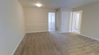 This single-floor space presents a straightforward, functional layout designed for comfortable everyday living. The main living area is generously sized, featuring wall-to-wall beige carpeting that adds warmth and softness underfoot. Neutral cream-toned walls throughout create a bright, cohesive atmosphere, complemented by white baseboards and trim that provide clean visual contrast.

A flush-mount ceiling light fixture illuminates the space evenly, contributing to the room's welcoming feel. A horizontal chair rail detail runs along one wall, adding a subtle architectural touch to the otherwise minimalist interior.

Multiple doorways visible from the main area lead to connecting rooms and spaces. A louvered closet door offers practical storage, while an open doorway reveals a tiled area beyond, suggesting a kitchen, bathroom, or utility space. Another partially visible doorway hints at further rooms within the layout.

Floor vents are positioned throughout, indicating a central HVAC system. The space is currently unoccupied, allowing the clean lines, neutral palette, and functional floor plan to take center stage. Overall, the interior offers a blank canvas well-suited for a variety of furniture arrangements and personal styles.