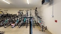This 620-square-foot bike room in Marlborough, MA, is a purpose-built facility designed to accommodate a large number of bicycles. The space features a clean, well-lit interior with bright fluorescent lighting and white walls, creating a safe and visible environment. The room is equipped with multiple bike storage solutions, including vertical and horizontal racks, maximizing the use of available space.

The facility boasts a concrete floor, ideal for durability and easy maintenance. Various types of bicycles are accommodated, from mountain bikes to road bikes and even children's bicycles, catering to diverse cycling needs. The storage systems appear to include both floor-mounted and wall-mounted options, allowing for flexible arrangement and efficient use of vertical space.

Access to the room is via a glass door, visible in some images, which likely provides security while allowing natural light to enter. The room also features modern amenities such as a ceiling-mounted projector or security camera, enhancing the space's functionality or security.

A fire extinguisher box is visible, indicating attention to safety measures. The overall layout of the room appears well-organized, with clear pathways between bike racks for easy access and navigation. This dedicated bike room offers a secure, convenient solution for bicycle storage, likely serving residents or employees of a larger complex or building.