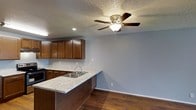 This single-floor residence offers a welcoming, open-concept layout that seamlessly connects the kitchen and living areas, creating a cohesive and functional living space. The kitchen is outfitted with rich, dark wood cabinetry that provides generous storage, complemented by light stone-patterned countertops that extend into a peninsula — ideal for casual dining or meal preparation. A double stainless steel sink anchors the peninsula, adding practicality to the space. Recessed lighting and a ceiling fan with a built-in light fixture ensure the area remains bright and well-ventilated throughout the day. The floors throughout are finished in warm, dark hardwood, lending a classic and durable character to the interior. Neutral light gray walls serve as a versatile backdrop, tying together the warm tones of the cabinetry and flooring. A textured ceiling adds a traditional architectural touch consistent throughout the space. The open flow between the kitchen and the adjoining living area enhances the overall sense of space, making this home well-suited for both everyday living and entertaining.