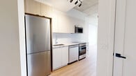 Located at 9450 Rue Meilleur in Montréal, this contemporary unit offers a well-planned single-floor layout comprising an open-concept kitchen and living area, one bathroom, and a private balcony. The living space is defined by warm light wood flooring, crisp white walls, and exposed concrete ceilings that lend an industrial-modern character throughout. Large black-framed floor-to-ceiling windows and a glass balcony door flood the space with natural light while providing sweeping views of the surrounding Montréal cityscape. A wall-mounted air conditioning unit ensures year-round comfort. The kitchen is efficiently designed with white cabinetry, a white tile backsplash, stainless steel appliances including a refrigerator, electric range, built-in microwave, and dishwasher, all finished with light countertops for a clean, cohesive look. The bathroom features a combined soaking tub and shower enclosure with a glass partition, set against bold dark granite-style wall tiles that contrast elegantly with the white fixtures and polished floor tiles. The private balcony, accessible through the living area, extends the living experience outdoors with open views over the neighbourhood. The overall ambiance is modern, bright, and functional.