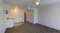 This single-floor unit features an open-concept layout that integrates the living area and kitchen into one shared space. The living area is finished with neutral-toned carpet, providing a soft underfoot feel, while the kitchen zone transitions to resilient tile-style flooring, clearly delineating the two functional areas. The kitchen is equipped with white cabinetry, a sink, and a full-size refrigerator, offering practical storage and workspace within a compact footprint. A ceiling fan with a built-in light fixture is centrally positioned overhead, supporting both air circulation and ambient lighting throughout the main space. The textured ceiling adds subtle visual depth to the interior. A central hallway, visible from the main living area, leads to additional rooms, including what appears to be a closet or storage space and a separate door providing further access. HVAC vents are integrated into the walls and ceiling, ensuring climate control throughout. The overall color palette is light and neutral, with white walls and trim contributing to a clean, understated aesthetic. The layout makes efficient use of the available space while maintaining clear separation between living and functional zones.
