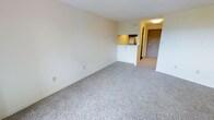 This single-floor unit offers a straightforward, functional layout centered around an open living area finished with neutral, light-toned carpet that extends throughout the main space. The walls are painted in a clean, off-white tone, creating a bright and airy atmosphere that enhances the sense of openness despite the unit's modest footprint.

A pass-through counter connects the compact kitchen to the living area, allowing for an open sightline between the two spaces. The kitchen features under-cabinet lighting and built-in range hood ventilation, providing practical utility in a condensed area. The warm-toned cabinetry and yellow-accented lighting add a subtle contrast to the otherwise neutral palette of the unit.

The entryway is visible from the living area, separated by a short corridor that leads to a solid entry door, offering a defined transition between the interior and exterior. An overhead light fixture in the hallway provides adequate illumination near the entrance. The overall layout is efficient, with a seamless flow between the living space, kitchen, and entryway, making the unit practical for everyday living.