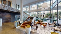 1900 Ocean Beach Club presents an architecturally striking lobby that immediately conveys a sense of grandeur and coastal sophistication. The centerpiece of the space is a dramatic double-height atrium framed by expansive floor-to-ceiling glass walls, flooding the interior with natural light and offering lush views of palm trees and the surrounding landscaping. A grand staircase with warm wood treads and sleek glass railings rises elegantly through the space, anchored by polished concrete floors that extend throughout.

The main lounge area is thoughtfully curated with an eclectic mix of modern and organic furnishings — including woven accent chairs, a tufted bench, sculptural side tables, and oversized decorative planters — all arranged atop a bold patterned area rug. Oversized pendant lighting adds warmth and drama overhead.

A dedicated reception desk clad in richly veined marble serves as the formal welcome point, complemented by a reclaimed wood ceiling that adds rustic warmth. Adjacent spaces include a private conference or meeting room featuring a long hardwood table, molded white chairs, a built-in aquarium, and a reclaimed wood ceiling — ideal for gatherings or events.

The entry vestibule offers seamless transparency with glass walls and doors, while a luggage cart signals attentive hospitality services. Together, these elements create a refined yet welcoming environment that balances natural materials with modern design.