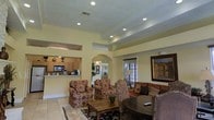 This single-floor space showcases a thoughtfully designed open layout that blends comfort with classic architectural detail. The living area is the heart of the space, featuring a prominent tray ceiling with recessed lighting that adds depth and a sense of elevation to the room. Warm, neutral-toned walls complement the light tile flooring, creating a bright and welcoming ambiance throughout.

A graceful arched doorway serves as a defining architectural element, connecting the living area to adjacent spaces and adding a touch of elegance to the overall design. The living room is generously appointed with plush, patterned armchairs and a rich upholstered sofa arranged around a solid wood coffee table and a round accent table, offering a comfortable and functional gathering space. A large framed artwork anchors one wall, adding a decorative focal point.

The kitchen is open to the living area via a pass-through counter, allowing natural sightlines between the two spaces. It features wood-toned cabinetry, modern appliances including a built-in microwave, and ample counter space. Natural light filters in through slatted blinds on the windows, enhancing the airy feel of the space. Overall, the layout and finishes combine to create a cohesive, inviting environment well-suited for both everyday living and entertaining.