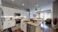 This single-story residence at Kinstead presents a thoughtfully designed open-concept floor plan finished with contemporary details throughout. The kitchen serves as a central focal point, showcasing crisp white cabinetry extending to the ceiling, a classic white subway tile backsplash, and a full suite of stainless steel appliances including a range, microwave, refrigerator, and dishwasher. A generously sized kitchen island with light quartz countertops provides additional workspace and seating, illuminated by industrial-style pendant lights overhead. Rich wood-tone flooring flows seamlessly from the kitchen into the dining and living areas, which benefit from natural light streaming through large windows with views of the surrounding landscape. The living space offers comfortable proportions with ceiling fan overhead.

The bedrooms are well-appointed with large windows that invite natural light and outdoor views, complemented by stylish curtain panels. The primary bathroom features wood-look flooring, a vanity with under-lit cabinetry, a lighted mirror, a glass-enclosed walk-in shower, and direct access to a walk-in closet with built-in shelving. An additional bathroom with a glass shower enclosure is also visible. Recessed lighting throughout enhances the clean, modern ambiance of the entire residence.