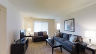 This single-floor space centers around a thoughtfully designed living room that combines comfort and functionality. The room is finished with neutral beige walls and smooth ceilings, creating a clean, understated backdrop. Carpeted flooring adds warmth underfoot and contributes to the overall cozy atmosphere.

The living area accommodates two dark charcoal-upholstered sofas accented with patterned throw pillows, arranged around a dark-finished rectangular coffee table. A matching media console along one wall houses a flat-screen television, keeping the entertainment setup organized and streamlined. A classic table lamp provides warm supplemental lighting alongside the room's natural light source.

Natural light enters through a large window dressed with vertical blinds, allowing flexible light control throughout the day. A secondary window is visible in the background, further brightening the space. A framed piece of wall art adds a decorative focal point, enhancing the room's polished yet approachable aesthetic. Overall, the living area presents a practical and welcoming environment with a cohesive, contemporary design sensibility.