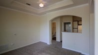 This single-story space showcases a thoughtfully designed interior with several notable architectural details. The main living area features a tray ceiling with crown molding, adding visual depth and a refined touch to the room. A ceiling fan is centrally mounted within the tray, providing both function and style. Neutral-toned carpet covers the floor, contributing to a warm and cohesive atmosphere throughout.

A prominent arched opening serves as a graceful transition point, connecting the living space to an adjacent hallway that leads to what appears to be a bathroom and walk-in closet. This architectural detail adds character and a sense of openness to the layout. On the opposite side, a partial-height wall separates the living area from the kitchen, which is equipped with white appliances including a refrigerator, stove, and microwave. This peninsula-style divider maintains visual connectivity between the two spaces while defining each area's function.

The walls are finished in a soft, neutral tone throughout, complementing the bright white trim and baseboards. The overall ambiance is clean, understated, and move-in ready, with classic design elements that provide a versatile backdrop for a variety of interior styles.