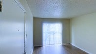This single-story space offers a clean and functional layout highlighted by warm hardwood-style flooring that runs throughout the main living area. A large sliding glass door serves as a focal point of the room, fitted with vertical blinds that allow for adjustable natural light and providing a direct view to an outdoor area beyond. The walls are painted in a neutral, soft tone that complements the flooring and contributes to a bright, open atmosphere. A textured popcorn ceiling adds a classic architectural detail overhead. Crisp white baseboards and door trim provide clean contrast against the walls, while a paneled entry door with hardware is visible to one side of the room. The overall layout is open and unobstructed, offering flexibility for various furniture arrangements. The abundance of natural light filtering through the sliding glass door creates an inviting and airy ambiance throughout the space.