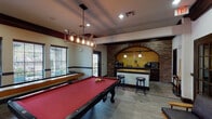Located in Houston, TX, this single-story game room is a well-appointed recreational space designed for both entertainment and casual gathering. The main area is anchored by a full-size pool table with a bold red felt surface, positioned beneath an industrial-style rectangular pendant light fitted with Edison bulbs. A shuffleboard table runs along the windowed wall, which offers generous natural light through multiple large windows with exterior views. A wall-mounted flat-screen television is positioned opposite the windows, adding a media element to the space.

Dark wood crown molding frames the ceiling throughout, lending a polished contrast to the light-toned walls and wood-look plank flooring. A kitchenette area serves as a standout architectural feature, framed by a dramatic full brick arched surround. Inside the arch, decorative yellow tile creates a warm backdrop for the counter space, which includes dark cabinetry, a sink, microwave, stainless steel appliances, a coffee station, and a granite-topped bar with bar stools. A chalkboard panel is mounted on the adjacent brick wall.

A lounge area provides a comfortable retreat, furnished with modern wooden armchairs and a round side table, accented by large, colorful abstract paintings that energize the walls. The overall ambiance balances rustic charm with contemporary design.