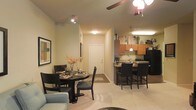 This single-floor residence offers a thoughtfully designed open-concept layout that seamlessly connects the main living areas. The dining area is a welcoming focal point, comfortably accommodating a round dining table and chairs beneath a ceiling fan with integrated lighting. Warm, neutral-toned carpet flows through the living and dining spaces, lending a cozy, comfortable ambiance.

The kitchen is efficiently designed and well-equipped, featuring warm wood-toned cabinetry that extends to the upper walls, providing generous storage space. A raised breakfast bar with seating for multiple guests bridges the kitchen and dining areas, making it ideal for casual dining and entertaining. Black appliances add a modern contrast to the cabinetry, and a pendant light above the bar enhances the kitchen's inviting atmosphere.

A clear sightline runs from the dining area down a hallway to the front entry door, emphasizing the home's open and airy feel. Recessed and track lighting throughout the space ensures each area is well-lit, while ceiling fans contribute to both comfort and style. The overall design strikes a balance between functionality and warmth, making this a practical and welcoming home.