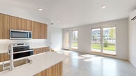 This contemporary two-story home is thoughtfully designed with clean lines and modern finishes throughout. The main level features an open-concept living and kitchen area anchored by polished concrete floors that lend an industrial-chic aesthetic. The kitchen is a standout space, equipped with warm wood-grain cabinetry, a white quartz countertop island with an integrated sink, stainless steel appliances including a refrigerator, range, and microwave, and a classic white subway tile backsplash. The adjacent living area is bathed in natural light, with multiple floor-to-ceiling windows and a glazed door that frames views of a lush green outdoor space. A clean, minimalist powder room with stone-toned tile flooring and brushed nickel fixtures completes the lower level, along with an attached garage featuring an automatic opener and ample room for vehicles. Upstairs, light-toned hardwood flooring flows through the bedrooms, each offering a bright, airy atmosphere with recessed lighting and generous closet space with built-in shelving. An additional sun-filled room on the upper floor provides flexible use as a bedroom or office. Throughout, crisp white walls and understated hardware reinforce a cohesive, modern design sensibility.