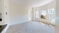 Located at 159 St Paul St in Brookline, Massachusetts, this single-floor unit offers a well-organized layout that balances comfort and functionality. The living area is bright and spacious, featuring soft wall-to-wall carpeting and a distinctive curved bay window that frames views of the surrounding neighborhood and allows generous natural light to fill the room. An adjacent bedroom continues the carpeted flooring and is notably well-equipped with dual built-in closets topped by upper cabinet storage, providing exceptional organizational capacity. A second carpeted room with two large double-hung windows and a wall-mounted heating unit adds further flexibility to the layout. The kitchen is efficient and neatly appointed, featuring white cabinetry, dark countertops, a white refrigerator, a stove, and a sink beneath overhead cabinets. Light marble-style tile flooring runs throughout the kitchen and entry area, creating a clean contrast to the carpeted living spaces. The bathroom is finished in crisp white subway tile, complete with a full bathtub and shower combination, chrome fixtures, a vanity with sink, and a mirrored medicine cabinet. The overall aesthetic throughout the unit is clean and understated, with neutral tones and white trim providing a bright, move-in ready environment.