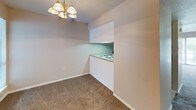 This single-floor unit is designed with a neutral, functional aesthetic throughout. The living room is a generously sized, carpet-floored space anchored by a large sliding glass door fitted with vertical blinds, allowing ample natural light and providing views of surrounding greenery. The dining area sits adjacent to the kitchen, illuminated by a classic brass chandelier, and connects to the kitchen via a convenient pass-through opening. The kitchen features coil-burner electric cooktop set into a laminate countertop, cabinetry with dark countertops, and a built-in range hood above, all within a compact, efficient layout.

The bedrooms are carpeted and well-sized, with neutral-toned walls and windows dressed with horizontal blinds for natural light control. One bedroom is notably well-equipped with two side-by-side bifold closets, offering generous storage capacity. Both bathrooms maintain a consistent design, featuring white cabinetry, louvered mirror cabinets, dark laminate countertops with white sinks, and tile flooring. One bathroom includes a shower-tub combination. Throughout the unit, white trim and soft beige tones create a clean, cohesive atmosphere.