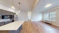 Located in Lacey, WA, this thoughtfully designed single-floor residence offers three bedrooms and two bathrooms within a well-appointed, contemporary layout. The open-concept kitchen and living area serves as the heart of the home, showcasing dark wood cabinetry with gold hardware, a white quartz countertop island, a classic subway tile backsplash, and stainless steel appliances including a full-size refrigerator and range. Elegant pendant lighting with brass accents illuminates the kitchen, adding warmth to the modern aesthetic. Warm-toned, hardwood-style flooring flows seamlessly throughout the entire residence, creating a cohesive and inviting atmosphere. The spacious living room is bathed in natural light through large windows and features a glass-paned exterior door, enhancing the bright and airy feel of the space. The primary bedroom is generously sized and connects to a well-appointed en-suite bathroom featuring a dual-sink vanity with dark wood cabinetry, quartz countertops, a large mirror with modern lighting, and an accessible walk-in shower with grab bars and a fold-down bench. Two closets provide ample storage, including a sizable walk-in closet with built-in shelving and hanging rods. The clean, white walls and modern finishes throughout give the entire residence a fresh, move-in ready appeal.