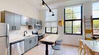 This single-floor space showcases a contemporary loft aesthetic defined by soaring ceilings and a series of large, black-framed multi-pane windows that flood the open interior with abundant natural light and frame views of surrounding greenery. The layout flows seamlessly from the kitchen through the dining area and into the living space, creating a cohesive and versatile environment throughout.

The kitchen is outfitted with a full suite of stainless steel appliances, including a refrigerator, electric range, built-in microwave, and dishwasher. Sleek gray cabinetry with matte black hardware pairs with white countertops and a black pull-down faucet over an undermount sink. Track lighting mounted to the ceiling provides focused task illumination. A butcher-block-topped island on casters with bar stools serves as a casual gathering and prep space.

The dining area features a simple white table and light wood chairs, while the living area accommodates a neutral sofa and additional seating beneath the dramatic floor-to-ceiling windows. Light hardwood flooring runs throughout, enhancing the bright, airy atmosphere.

The bathroom is cleanly finished with large-format wall tile, a frameless sliding glass shower enclosure with a pebble-tile floor, and matte black shelving for storage.