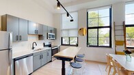 This single-floor space showcases a contemporary loft aesthetic defined by soaring ceilings and a series of large, black-framed multi-pane windows that flood the open interior with abundant natural light and frame views of surrounding greenery. The layout flows seamlessly from the kitchen through the dining area and into the living space, creating a cohesive and versatile environment throughout.

The kitchen is outfitted with a full suite of stainless steel appliances, including a refrigerator, electric range, built-in microwave, and dishwasher. Sleek gray cabinetry with matte black hardware pairs with white countertops and a black pull-down faucet over an undermount sink. Track lighting mounted to the ceiling provides focused task illumination. A butcher-block-topped island on casters with bar stools serves as a casual gathering and prep space.

The dining area features a simple white table and light wood chairs, while the living area accommodates a neutral sofa and additional seating beneath the dramatic floor-to-ceiling windows. Light hardwood flooring runs throughout, enhancing the bright, airy atmosphere.

The bathroom is cleanly finished with large-format wall tile, a frameless sliding glass shower enclosure with a pebble-tile floor, and matte black shelving for storage.