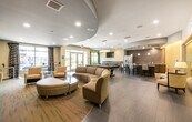This single-floor amenity space is designed with a contemporary aesthetic, combining stylish finishes and thoughtfully arranged zones to create a dynamic communal environment. The entrance foyer greets visitors with large glass doors that open directly to an outdoor pool and patio area, flanked by tall windows dressed with dark drape panels and accented by potted greenery, allowing abundant natural light to flow inward.

The main lounge area is anchored by a generously sized curved sectional sofa in warm earth tones, paired with a two-tone oval coffee table. A distinctive curved ceiling detail in dark gray defines this social hub, which also features a built-in media wall with dark wood cabinetry housing a large flat-screen television and a sleek soundbar. Patterned armchairs, a bistro-height table with bar stools, and framed abstract artwork contribute to the sophisticated atmosphere.

Adjacent to the lounge sits a billiards table set atop hardwood-style flooring, offering a dedicated recreation zone. A well-appointed kitchenette nearby features quartz countertops, dark wood cabinetry, stainless steel appliances including a full refrigerator, and pendant lighting overhead.

A separate corridor area provides additional seating with glass-walled conference or flex rooms visible alongside curated artwork and recessed lighting throughout.