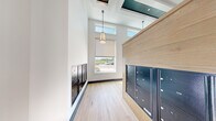This dedicated mail room is a thoughtfully designed communal space that balances practicality with modern aesthetics. The room is organized around multiple banks of sleek, matte black mailbox units that line both the perimeter walls and a central freestanding partition clad in light oak wood paneling — creating a clean, contemporary visual contrast between warm wood tones and dark metal hardware. The light oak wood-look flooring runs throughout, lending a cohesive and polished feel to the space. Soaring ceiling heights amplify the sense of openness, and clerestory windows positioned near the top of the walls allow ample natural light to filter in, brightening the interior without sacrificing privacy. A central pendant light fixture with a classic drum shade hangs from the ceiling, providing warm, focused illumination. One end of the room features a full-height window with a roller shade, offering an exterior view and additional daylighting. Decorative patterned wallcovering accents the upper walls in one section of the room, adding visual interest and texture. The overall ambiance is clean, modern, and functional — designed to serve a multi-resident building efficiently while maintaining an elevated, welcoming environment.