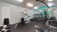 This fitness center is a thoughtfully designed exercise space finished with a combination of teal and neutral gray walls that create an energetic yet focused atmosphere. The vaulted ceiling adds a sense of openness to the room, which is brightly illuminated by flush-mounted fluorescent light fixtures overhead. Dark rubber flooring covers the entire space, providing a durable, safety-conscious surface appropriate for physical activity.

The cardio area features multiple treadmills and an elliptical trainer, positioned along one side of the room. A large mirrored wall panel runs along this section, visually expanding the space and allowing users to monitor their form during workouts. Strength training equipment includes a multi-function cable machine, a leg press or extension station, a dumbbell rack with free weights, and several workout benches. A wall-mounted flat-screen television is installed near the entrance, offering entertainment during exercise sessions.

Climate control is provided by a wall-mounted HVAC unit, and a fire extinguisher is conveniently accessible near the entry door. A motivational "Fitness" wall decal adds a finishing touch to the space. The room is accessed through a single entry door and is designated exclusively as a fitness center.
