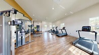 Located in Fort Pierce, Florida, this fitness center at 2935 Ashland Cir offers a clean, modern space designed to support a well-rounded workout experience. The facility is comprised of a spacious exercise room and a connecting hallway, all situated on a single level.

The exercise room is bright and welcoming, featuring crisp white walls, recessed lighting, and warm wood-look flooring throughout. Large multi-pane windows line the walls, flooding the space with abundant natural light and providing views of the surrounding outdoor greenery. The high ceilings contribute to an open, airy atmosphere that enhances the sense of space.

The room is well-appointed with a diverse selection of fitness equipment, including treadmills, elliptical trainers, a rowing machine, a stair climber, and a cable/functional training machine. A dedicated free weights station features a tiered dumbbell rack, complemented by exercise balls and resistance equipment for varied training options. A large floor-to-ceiling mirror along one wall expands the visual space and supports form-checking during workouts. A small utility counter area provides additional convenience.

French doors at one end of the exercise room open to an outdoor area, offering easy access and enhanced ventilation. The hallway connects to additional rooms with multiple doors, providing utility and organization to the overall layout. The facility presents a polished, functional environment suitable for a broad range of fitness activities.