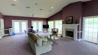 Located at 501 Fairways Dr in Vicksburg, Mississippi, this single-story home offers a thoughtfully designed layout centered around a generous, open living area. The room immediately draws attention with its soaring vaulted ceilings that create an expansive, airy atmosphere throughout the space. Rich plum-toned walls provide a bold, warm contrast to the crisp white ceiling and trim, lending the interior a distinctive and inviting character.

The living area is anchored by a classically styled white-manteled fireplace with a tile surround, serving as a prominent focal point. Soft carpeting extends across the floor, adding warmth and comfort underfoot. Recessed lighting and a ceiling fan are integrated into the vaulted ceiling, ensuring both practical illumination and comfortable airflow.

Natural light flows generously into the room through multiple large windows and French doors, which line the walls and offer views to the outdoors. This abundance of glazing creates a bright, open feel that connects the interior with the surrounding environment.

The space comfortably accommodates distinct living and dining areas, with room for generous seating arrangements, casual dining furniture, and accent pieces. The overall ambiance is one of comfortable, unpretentious elegance — a welcoming home well-suited for both everyday living and entertaining.