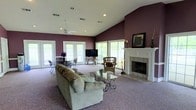 Located at 501 Fairways Dr in Vicksburg, Mississippi, this single-story home offers a thoughtfully designed layout centered around a generous, open living area. The room immediately draws attention with its soaring vaulted ceilings that create an expansive, airy atmosphere throughout the space. Rich plum-toned walls provide a bold, warm contrast to the crisp white ceiling and trim, lending the interior a distinctive and inviting character.

The living area is anchored by a classically styled white-manteled fireplace with a tile surround, serving as a prominent focal point. Soft carpeting extends across the floor, adding warmth and comfort underfoot. Recessed lighting and a ceiling fan are integrated into the vaulted ceiling, ensuring both practical illumination and comfortable airflow.

Natural light flows generously into the room through multiple large windows and French doors, which line the walls and offer views to the outdoors. This abundance of glazing creates a bright, open feel that connects the interior with the surrounding environment.

The space comfortably accommodates distinct living and dining areas, with room for generous seating arrangements, casual dining furniture, and accent pieces. The overall ambiance is one of comfortable, unpretentious elegance — a welcoming home well-suited for both everyday living and entertaining.